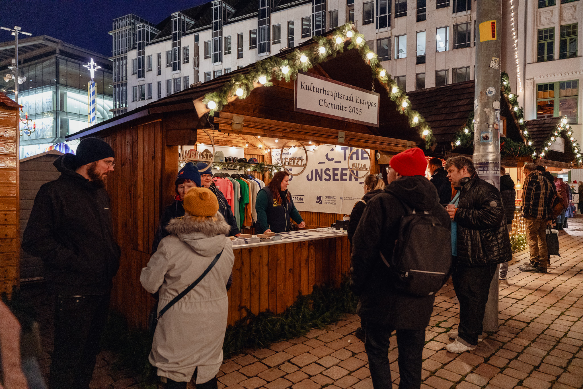 People gather around a festive wooden stall decorated with fairy lights at a night market in Chemnitz. The sign at the top reads "Chemnitz European Capital of Culture 2025". Some browse through the offerings while others chat nearby. Buildings can be seen in the background.